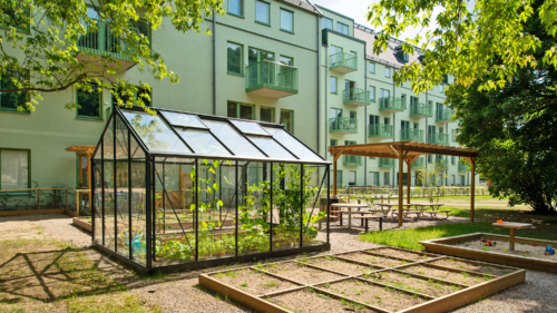 En grön gemensam trädgård framför ett modernt lägenhetshus med ett stort växthus i mitten, omgiven av upphöjda odlingsbäddar och sittplatser under en pergola.